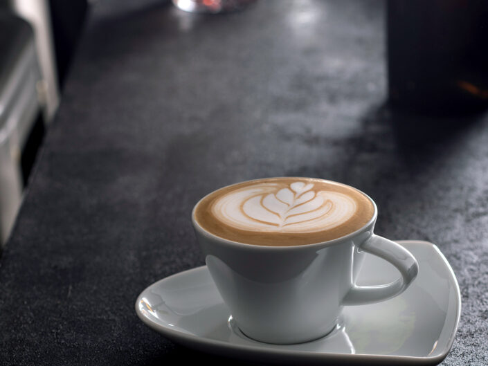 Coffee Art, Cappuccino mit Blume auf Theke, Eiskaffee im Hintergrund, weiße Tasse, dreieckige Untertasse, Barista