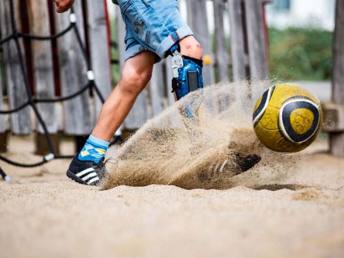 Junge mit Orthese am Bein spielt Fußball im Sand